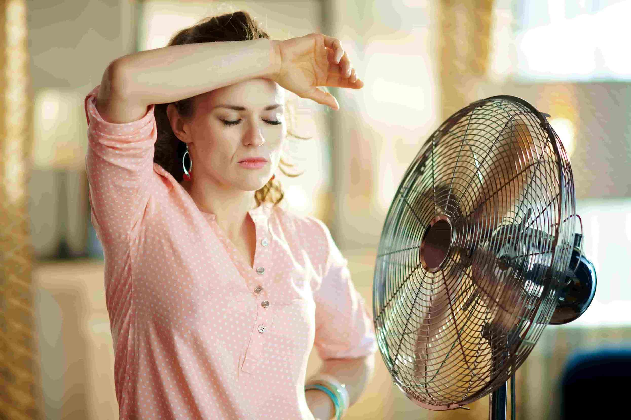 Exhausted woman facing in front of a stand fan - triggers for hot flashes